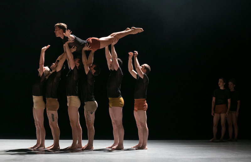 Dancers stand in a straight line holding another dancer above their heads. 